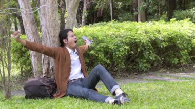 Asyalı adam akıllı telefon kullanır ve bahçe parkında bir ağacın altında oturur. Yavaş çekim VDO yaşam tarzını açık havada kavra.