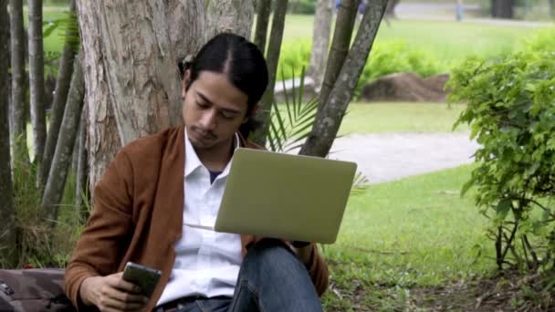 Hombre Asiático Utiliza Teléfono Inteligente Computadora Portátil Sienta Debajo Árbol — Vídeos de Stock