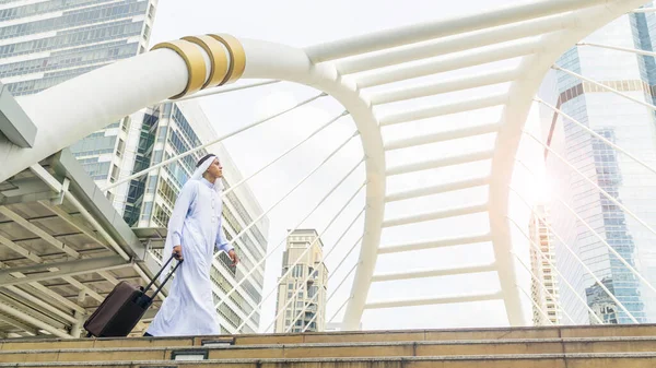 Business arab traveler saudi man carrying a suitcase and walk in the business landmark city with the modern steel structure and exterior wall modern building