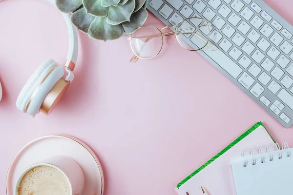 Flat lay of office feminine desk workspace with cup of coffee, succulent, laptop, empty notebook, headphones, keyboard, and golden eyeglasses on pink background. Top view. Copy space