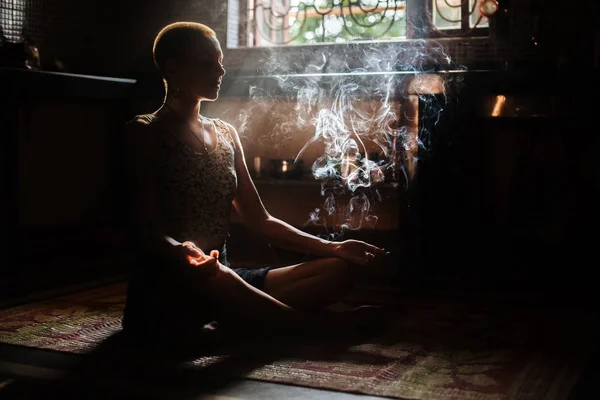 A girl in a meditation pose in the rays of the sun from a window in the smoke from a candle