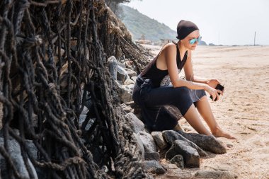 Taşlar kumlu bir plaj üzerinde oturan ve kulaklıklar müzik dinlemek gözlük kel spor şık genç kızla