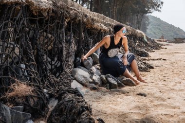 Taşlar kumlu bir plaj üzerinde oturan ve kulaklıklar müzik dinlemek gözlük kel spor şık genç kızla