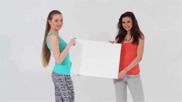 Due ragazze attraenti sorridenti in leggins sportivi e camicie guardando la fotocamera che tiene un banner bianco in bianco — Video Stock