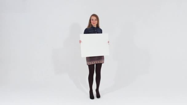 Sonriente chica atractiva en vestido marrón caminando y mirando a la cámara sosteniendo una pancarta blanca en blanco — Vídeo de stock