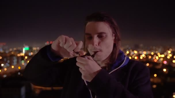 Молодой человек с деревянной трубкой зажигает табак на фоне ночного города — стоковое видео