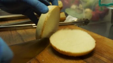 Ellerinde eldiven yarım hamburger çörek içinde kesin bir ahşap tahta üzerinde bir mutfak bıçağı ile