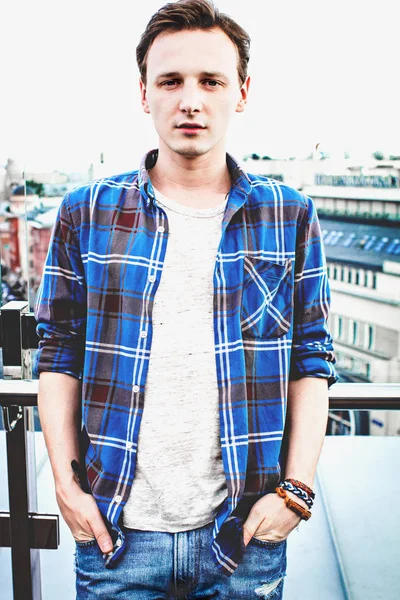 Cute trendy guy outdoor. Stylish man on the roof. Fashion total look: blue jeans, grey t-shirt and grunge shirt. Moscow background