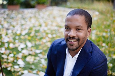 Şehir parkında oturan formal giyim gülümseyen kendine güvenen genç Afrikalı-Amerikalı işadamı portresi. Arka plan bulanık. Yatay.