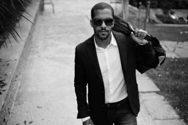 Formal giyim güneş gözlüğü ve Şehir Parkı, seyahat çantası ile yürüme mutlu kendine güvenen genç Afrikalı-Amerikalı işadamı portresi. Arka plan bulanık.