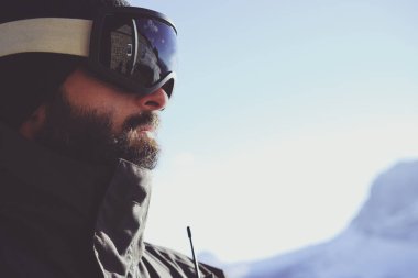 Sakallı genç yakın çekim portre sunglass maskesini, dağlar ve mavi gökyüzü arka plan üzerinde kayak merkezi snowboarded. Arka plan bulanık. Yatay