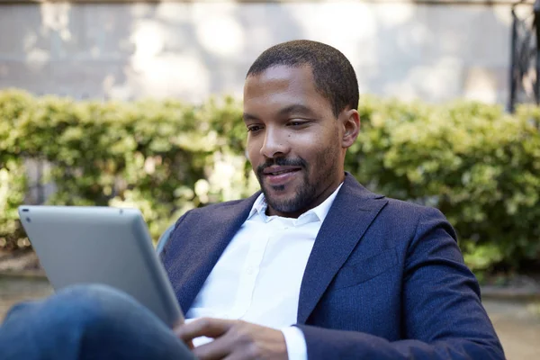 Empresário africano americano sorridente em roupas informais que trabalham na rua ensolarada em almofada de toque eletrônico, verificando e-mail usando dispositivo eletrônico habilitado para a Internet, chamando amigos através de redes sociais . — Fotografia de Stock