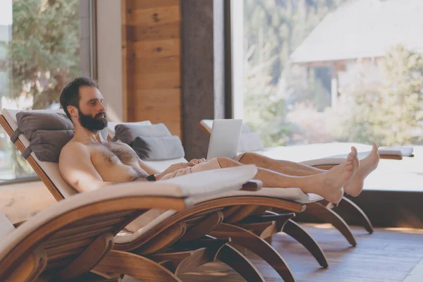 Handsome bearded Man using laptop computer while resting in spa hotel. Man relaxing after hard day at spa salon.Sunligth wooden interior