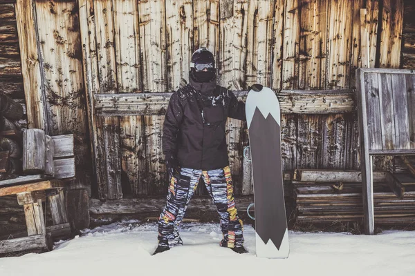 Snowboarder con chaqueta negra, máscara con snowboard cerrar auténtica casa de madera. Horizontal . — Foto de Stock