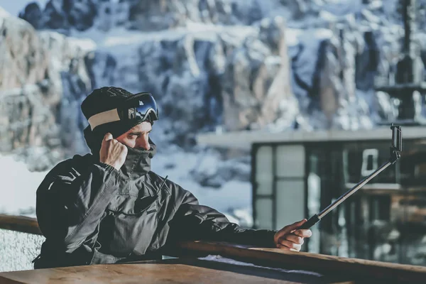 Barbudo jovem snowboard em máscara de vidro solar, na estância de esqui no fundo das montanhas e céu azul.Homem fazendo foto selfie em seu telefone inteligente. Horizontal.Blurred fundo . — Fotografia de Stock