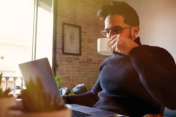 Bonito homem barbudo bonito usando computador portátil móvel enquanto bebe café na sala de estar em casa. Homem usando gadget eletrônico. Fundo desfocado . — Fotografia de Stock