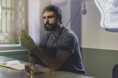 Genç kulaklık ile kafede oturan adam sakallı. Smartphone elele ve dijital müzik dinleme erkek. Notlar not defteri ve dizüstü bilgisayar, adam. Yatay. Arka plan bulanık.