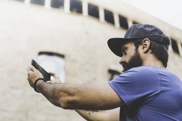 Hombre musculoso barbudo con gorra negra y haciendo selfie con su propio smartphone. Hombre caminando por la calle y usando móviles. Fondo borroso. Horizontal . —  Fotos de Stock