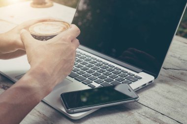 Yakın plan adam elinde akıllı telefon ve dizüstü bilgisayarla bir fincan kahve tutuyor. Rahatla konsept.