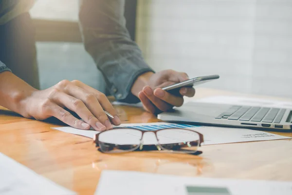 business man holding smartphone and writing financial paper work and laptop on wooden desk indoor office with morning light. Vintage filter effect.