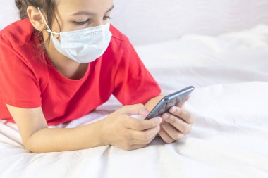 Uzaktan eğitim, online eğitim. Maskeli bir kız, okul ya da oyunlar için cep telefonu kullanıyor. Cep telefonuyla yatakta yatmak. Evde karantina.