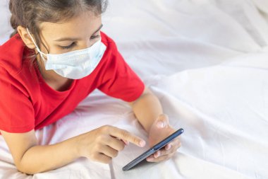 Distance schooling, learning online education. Girl wearing mask, using mobile for school or games. Laying on bed with mobile. Quarantine at home. clipart