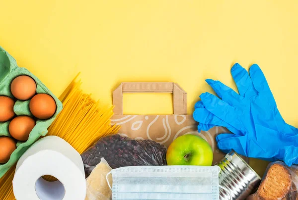 Food for quarantine isolation period flat lay on yellow background with copy space. Eggs, pasta, beans, toilet paper, apple and some seleals. Crisis food supplies. Delivery, donation concept.