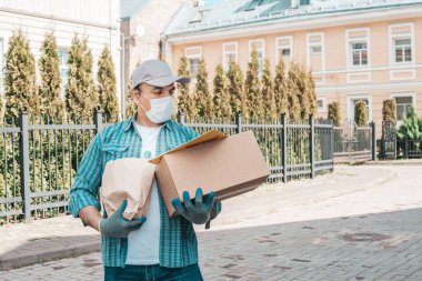 Koruyucu maskeli ve tıbbi eldivenli kurye paket yemek getiriyor. Teslimat servisi karantina altında. Karton kutu ve paket..