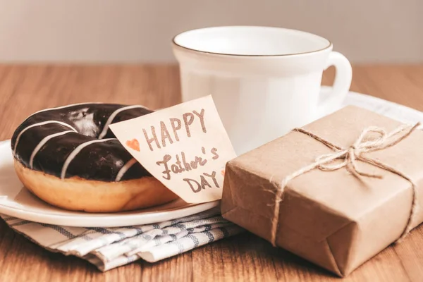 Gift box with a tag, cup of coffee or tea and chocolate donut. Fathers day or birthday concept.