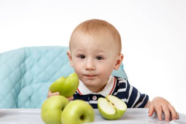Neşeli küçük çocuk çocuk çocuk koltuğunda oturuyor ve beyaz arka planda yeşil elma yiyor. Taze meyveler çocuklar için çok yararlıdır. Zeki yetişkinler iyi beslenme uzmanlarıdır..