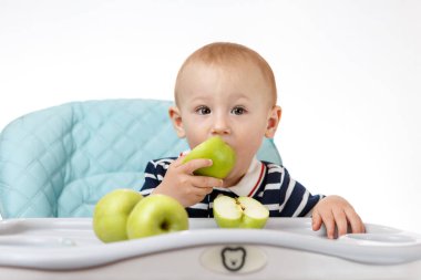 Neşeli küçük çocuk çocuk çocuk koltuğunda oturuyor ve beyaz arka planda yeşil elma yiyor. Taze meyveler çocuklar için çok yararlıdır. Zeki yetişkinler iyi beslenme uzmanlarıdır..