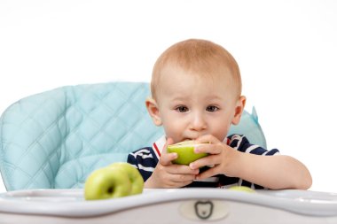 Neşeli küçük çocuk çocuk çocuk koltuğunda oturuyor ve beyaz arka planda yeşil elma yiyor. Taze meyveler çocuklar için çok yararlıdır. Zeki yetişkinler iyi beslenme uzmanlarıdır..