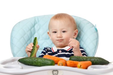 Neşeli küçük çocuk çocuk çocuk koltuğunda oturuyor ve beyaz arka planda yeşil bir salatalık yiyor. Taze sebzeler çocuklar için çok yararlıdır. Zeki yetişkinler iyi beslenme uzmanlarıdır..