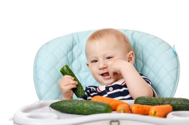 Neşeli küçük çocuk çocuk çocuk koltuğunda oturuyor ve beyaz arka planda yeşil bir salatalık yiyor. Taze sebzeler çocuklar için çok yararlıdır. Zeki yetişkinler iyi beslenme uzmanlarıdır..