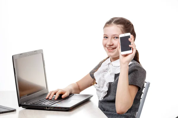 Linda colegiala sentada en un ordenador portátil y hablando por teléfono sobre un fondo blanco — Foto de Stock