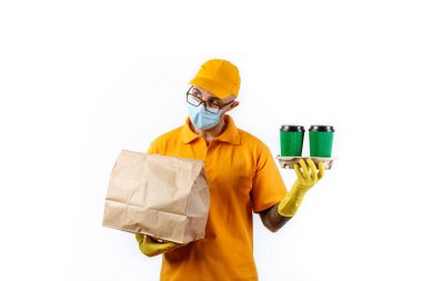 Tıbbi maskeli ve eldivenli kurye servisinden dikkatli genç bir adam bir fincan kahve ve beyaz arka planda bir paket tutuyor.