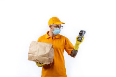 Tıbbi maskeli ve eldivenli bir kurye servisinden gelen ihtiyatlı genç adam nakit ödemeler için bir terminal ve beyaz arka planda bir paket tutuyor.
