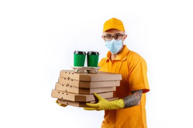 Tıbbi maskeli ve eldivenli kurye servisinden ihtiyatlı genç bir adam ellerinde beyaz bir arka planda kutu pizza ve kahve tutuyor.