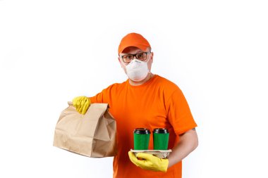 Tıbbi maskeli ve eldivenli kurye servisinden dikkatli genç bir adam bir fincan kahve ve beyaz arka planda bir paket tutuyor.