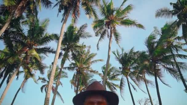 Młody człowiek aktywny w kapeluszu z wąsami w żółte okulary wśród drzew kokosowych wysyła pozdrowienia z kamerą, zdejmując kapelusz, wygłupiać się w aparacie. Pionierem w tropikalnej wyspie. Podróży — Wideo stockowe