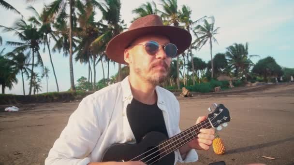 Un joven activo con un sombrero con bigote en gafas amarillas en la playa del océano juega en el ukelele entre las palmas de coco, tontos alrededor en cámara. Concepto de viaje — Vídeos de Stock