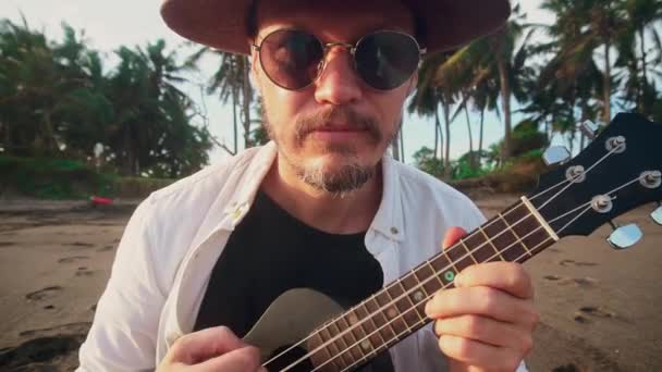 Um jovem ativo com um chapéu com um bigode em óculos amarelos na praia do oceano joga no ukulele entre as palmeiras de coco, tolos ao redor na câmera. Conceito de viagem — Vídeo de Stock