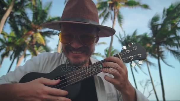 Um jovem homem ativo com um chapéu com um bigode em óculos amarelos joga no ukulele entre as palmeiras de coco, tolos em torno da câmera. Conceito de viagem — Vídeo de Stock