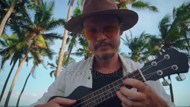 Een actieve jongeman met een hoed met een snor in gele glazen speelt op de ukulele onder de kokospalmen, dwazen rond op camera. Travel Concept — Stockvideo