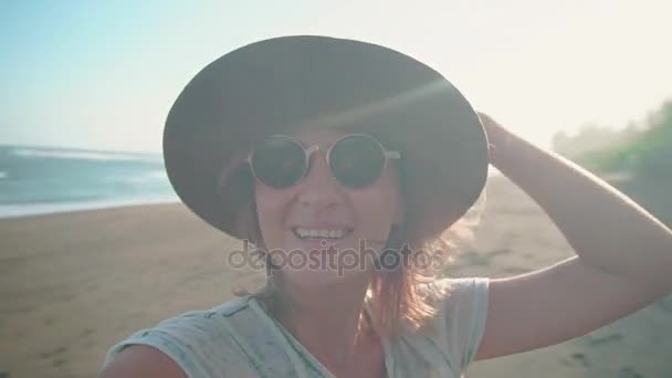 Uma jovem mulher com chapéu à luz do sol na praia do oceano sorri na câmera desfruta de novas aventuras, férias e viagens. Retrato de um turista em uma praia com palmeiras no fundo — Vídeo de Stock