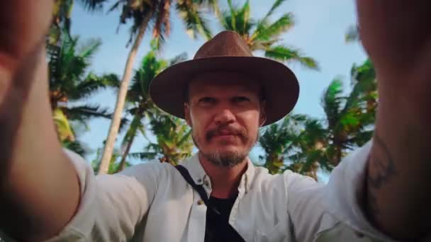 Un giovane uomo con un cappello con i baffi tra le palme da cocco tiene la fotocamera o il telefono tra le mani, si spara e invia saluti nella fotocamera — Video Stock