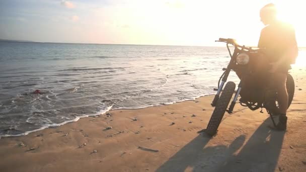 Motociclista começando a dirigir sua moto na praia durante o pôr do sol, câmera lenta — Vídeo de Stock