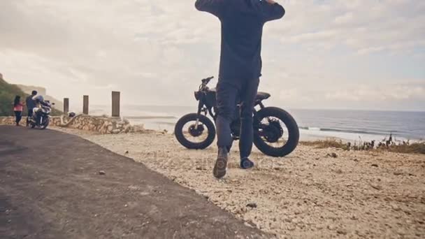 Un joven va a su motocicleta, se sienta en ella y se pone un casco para ir en un viaje a través del océano. Movimiento lento — Vídeos de Stock