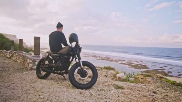 El motociclista se sienta en su motocicleta y admira una hermosa vista de las montañas y el océano, un alto en el camino — Vídeos de Stock