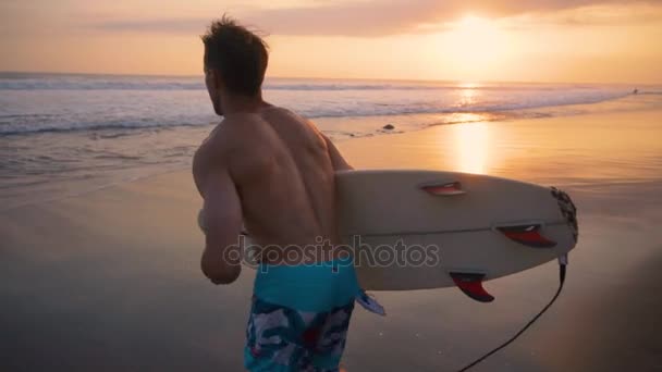 Young surfer holding board, running to go in the water at amazing golden light sunset. Happy man enjoying summer evening and seaside vacation activities. Shot taken by a handheld gimbal in 60FPS — Stock Video